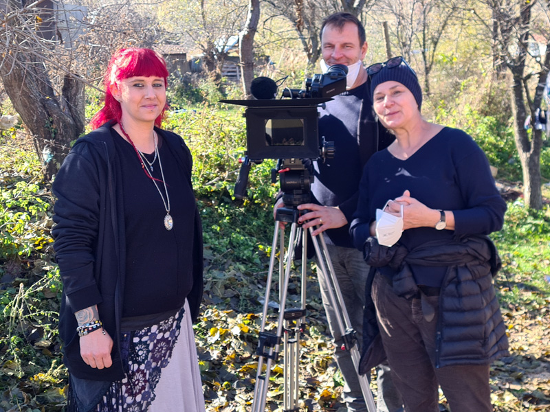 Zwei Frauen und ein Mann stehen in einem Garten. Mann steht hinter der Kamera auf Stativ.